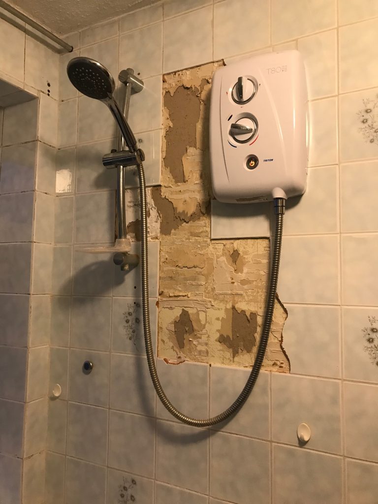 The old electric shower with some blue and floral tiles still visible around it, but a few tiles already removed. The whole situation looks very sad, not helped by the poor lighting conditions for the photo.