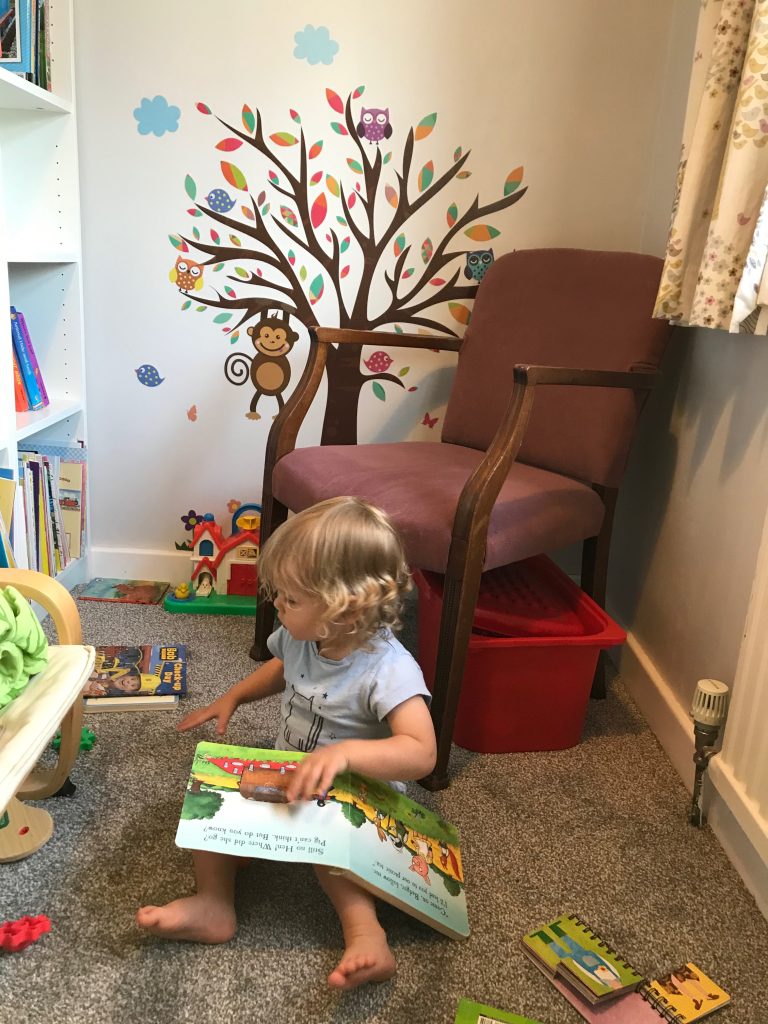 A two year old child sat on the floor of a newly decorated bedroom looking at books. The carpet on the floor looks very new and on the grey wall is a sticker picture of a large tree with colourful leaves and moneys and owls in it.
