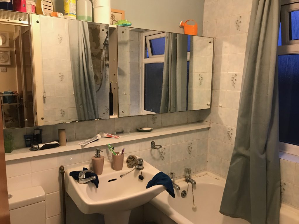 A view of a very 80s style bathroom with pale blue floral tiles and a sad shower curtain in shot. You can see the sink and taps end of the bath and also the shelf that runs along that wall of the bathroom and also teh mirrored cabinets. Poor lighting makes not look even more miserable than it was.
