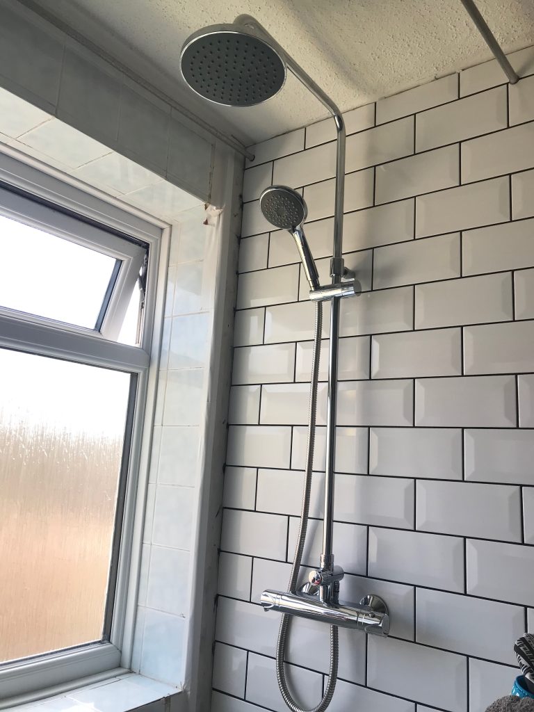 A wall tiled in white metro tiles with black grout and showing a shiny new shower with both a handheld he'd and a rainfall head. The adjacent wall still has the 1980s style blue tiles on it.