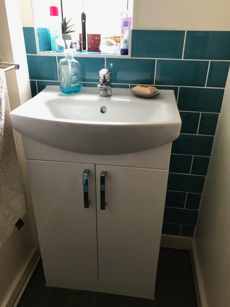 A modern looking washbasin. White with a white built in cupboard underneath it. Around teh sink are some petrol blue metro style tiles.