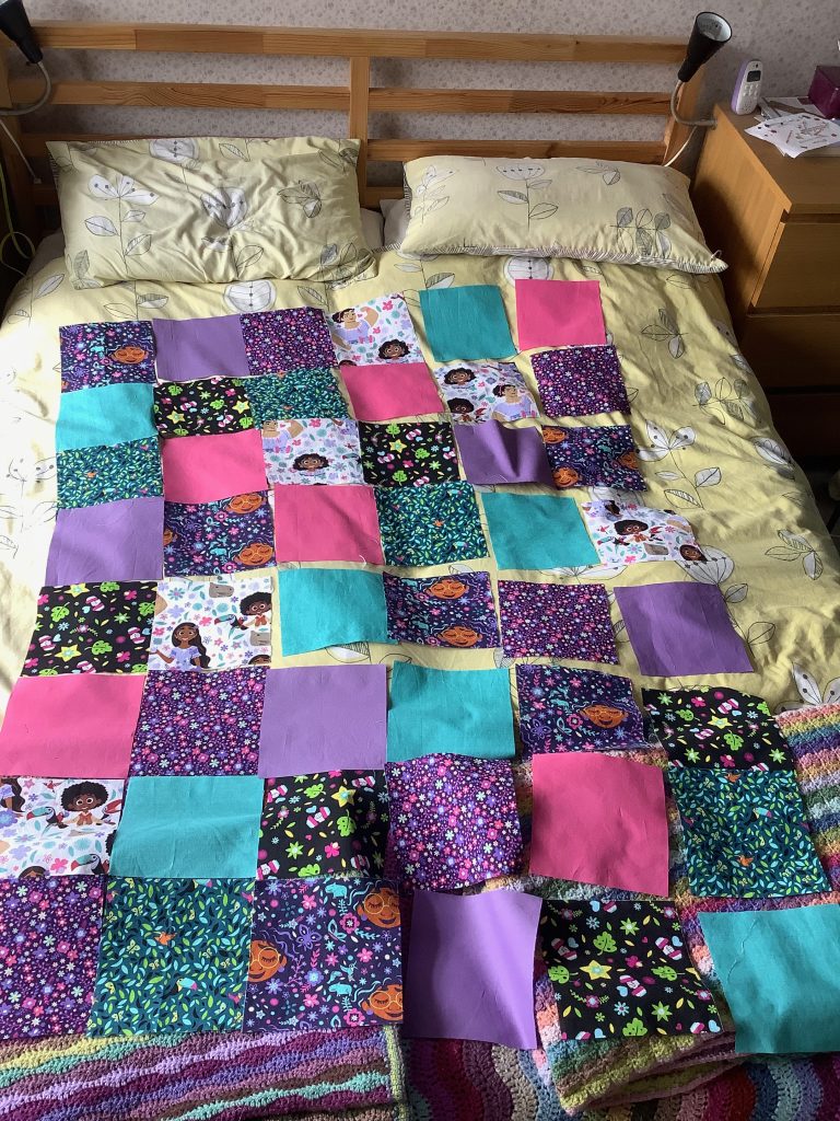 Fabric squares laid out on a double bed. Sevela of teh squares have an Encanto theme to them, whilst the others are coordinating plain coloured blocks.