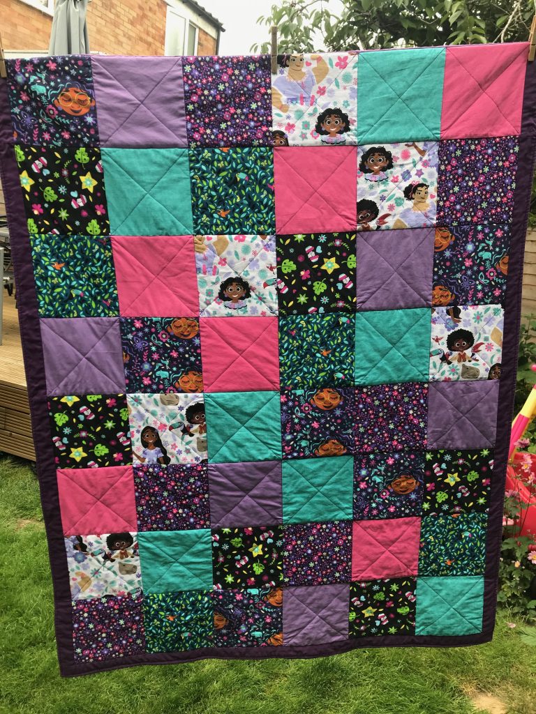 A completed Disney Encanto themed quilt hung on the washing line. It has a purple boarder all around.