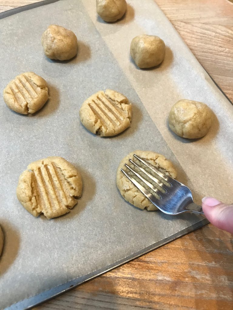 A baking tray of round mounds of biscuit dough with a fork pressing one of them down before they go in the oven
