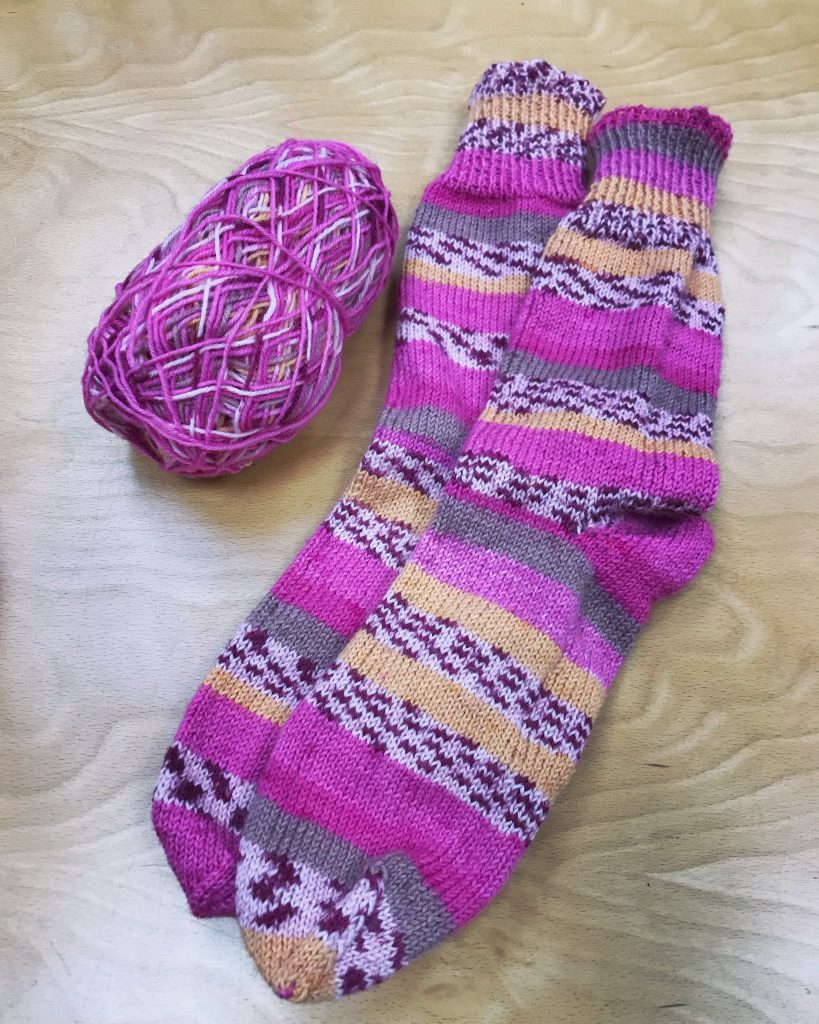 A completed pair of hand knitted socks. They are striped, but not matching with a main bright pink colour and yellow, brown and black and white patterned bits. Next to them is the remains of a ball of yarn.