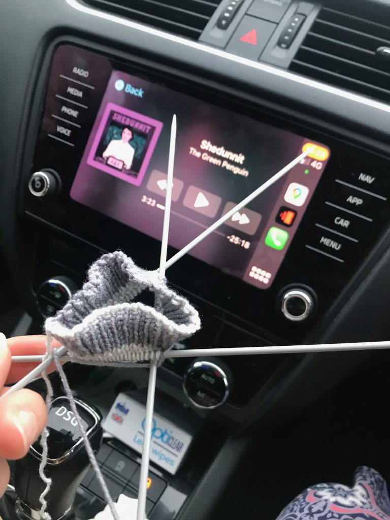 The start of a hand knitted sock on three double pointed needles. It can be seen that this is being knit in a car and in the background is the Shedunnit podcast playing on my car's audio screen.
